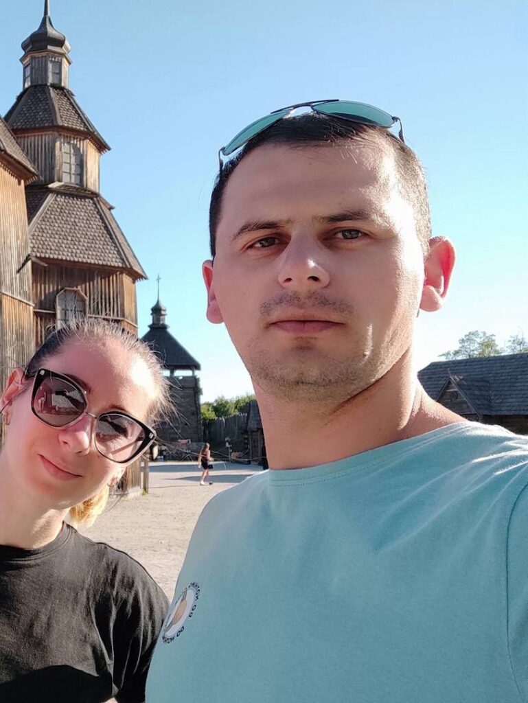 Alexey sammen med sin kone tager en selfie foran en historisk trækirke på en solrig dag.