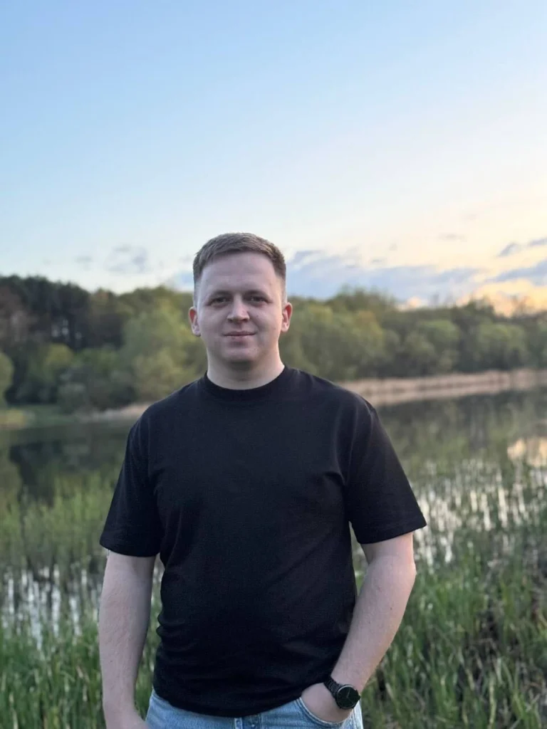 Portræt af Maksym Yurchenko stående udendørs ved en sø ved solnedgang.